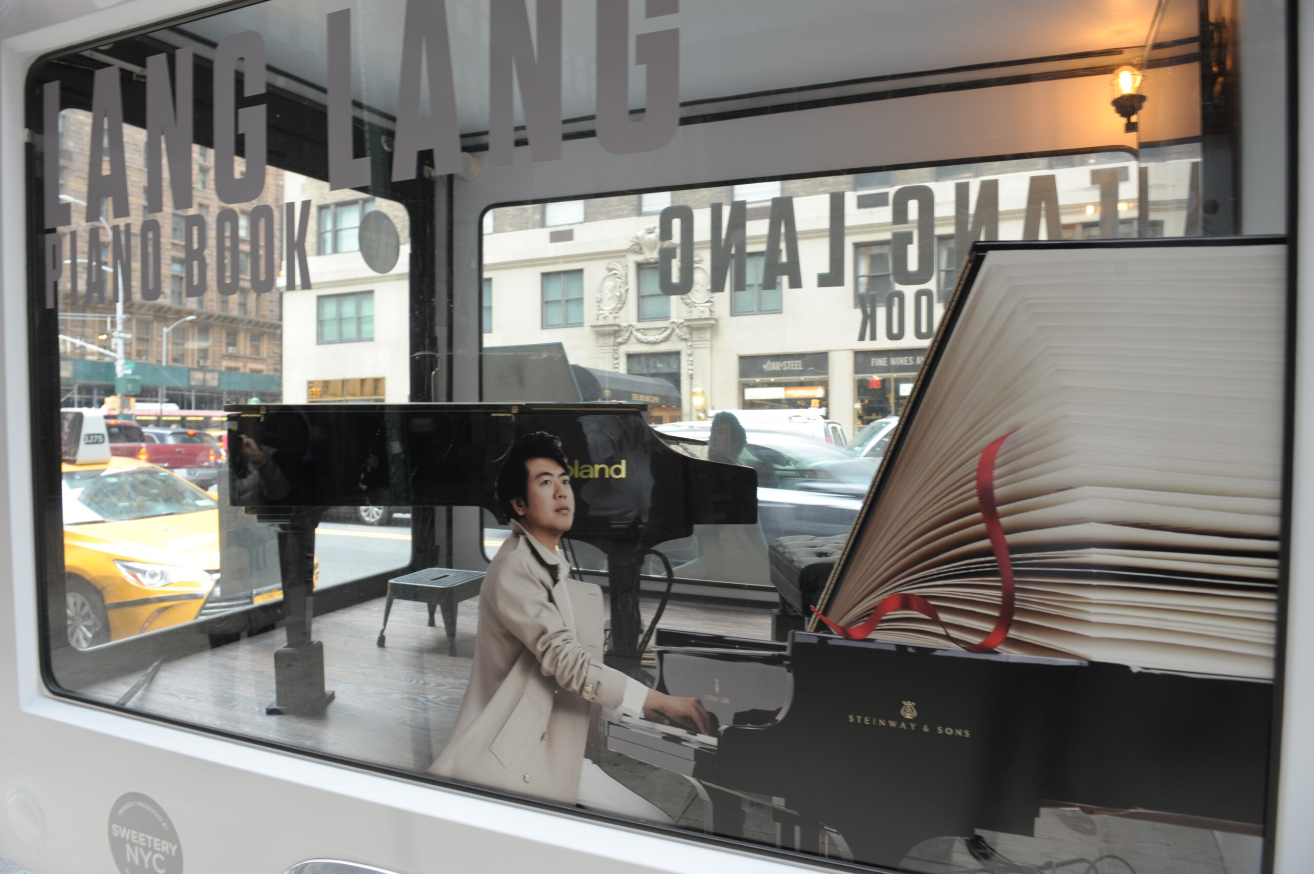 Lang Lang Piano Book glass box truck with a grand piano and oversized open book display inside a street activation.