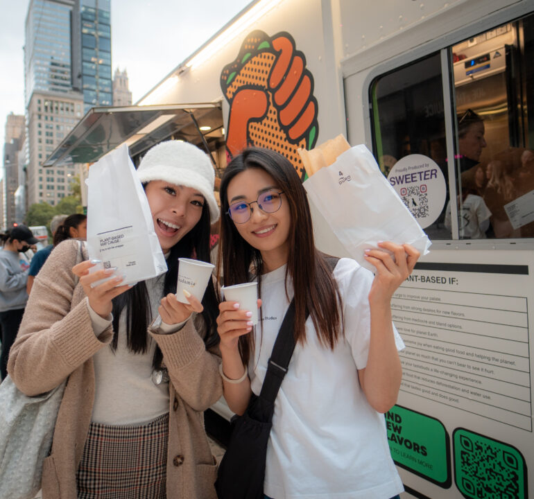 Two customers holding Grow Plant Based sample bags and drinks in front of the branded food truck activation.