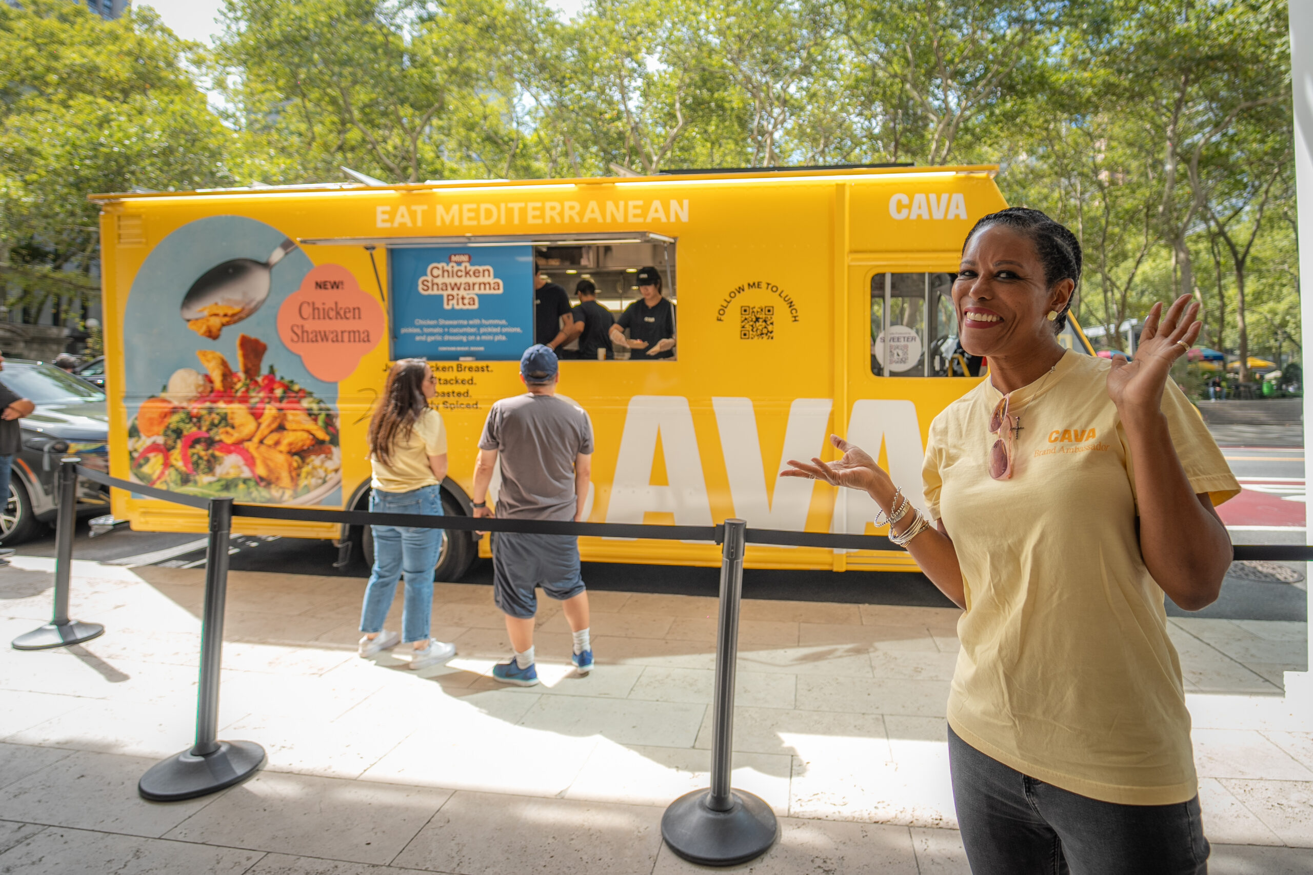 CAVA brand ambassador posing in front of a yellow branded food truck with customers ordering chicken shawarma samples.